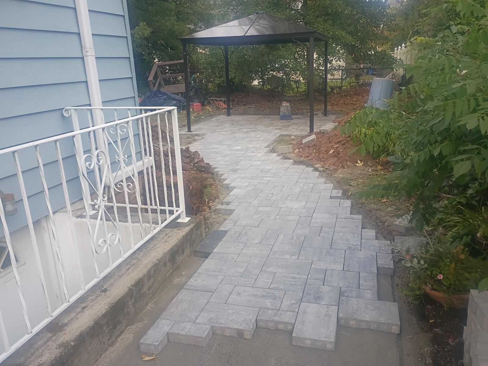 Paver pathway leading to a gazebo, alongside a blue house and white railing.