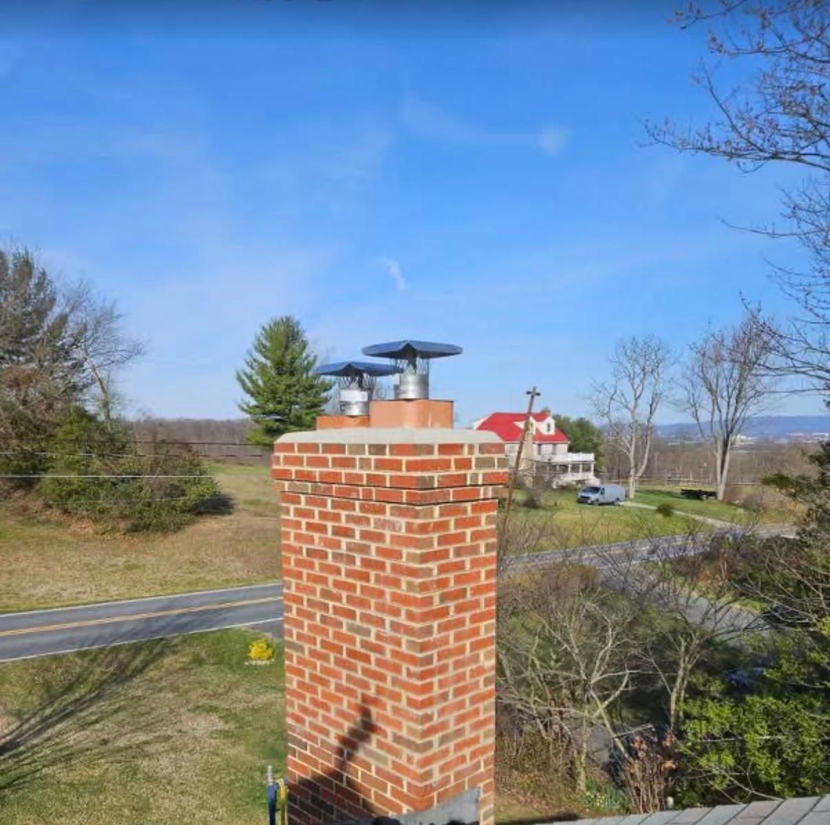Brick chimney with two metal caps, overlooking a road and a distant house with a red roof under a blue sky.