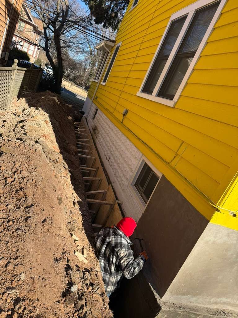 A person works in a narrow, muddy trench next to a yellow house. Wooden steps lead down.