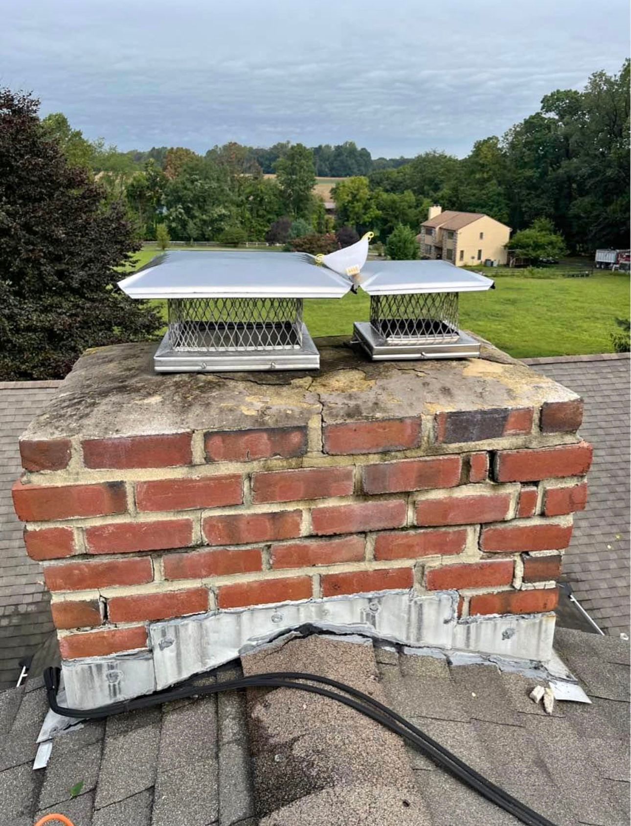 Chimney with two metal caps atop red bricks, on a roof with a green landscape backdrop.
