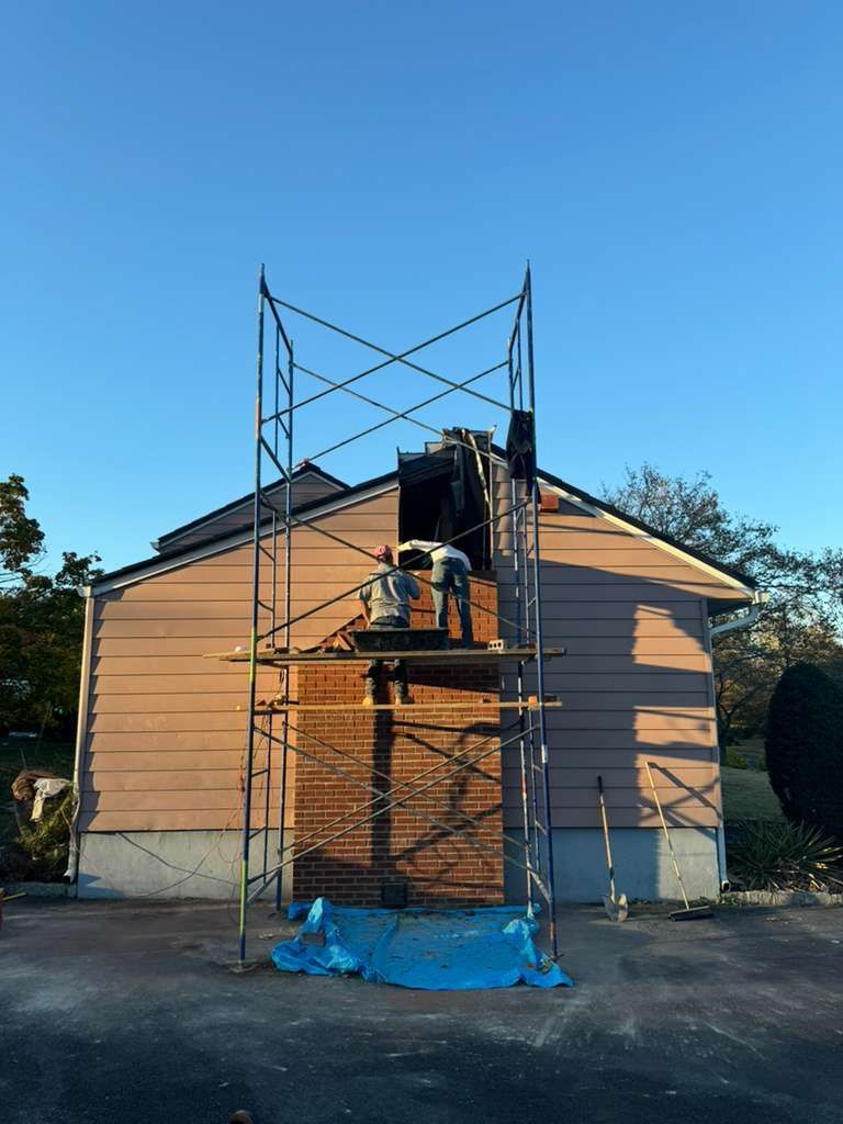 Two workers on scaffolding repair a brick chimney on a house, under a clear blue sky.