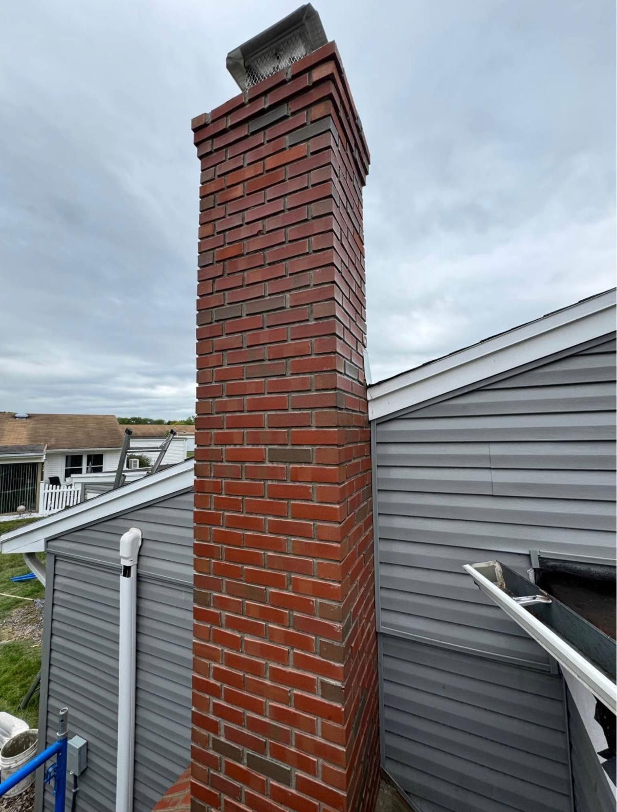 Brick chimney extending upwards from a gray-sided house against a cloudy sky.