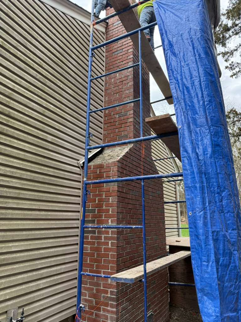 Brick chimney with scaffolding, being worked on near a house with beige siding. Blue tarp covers the side.