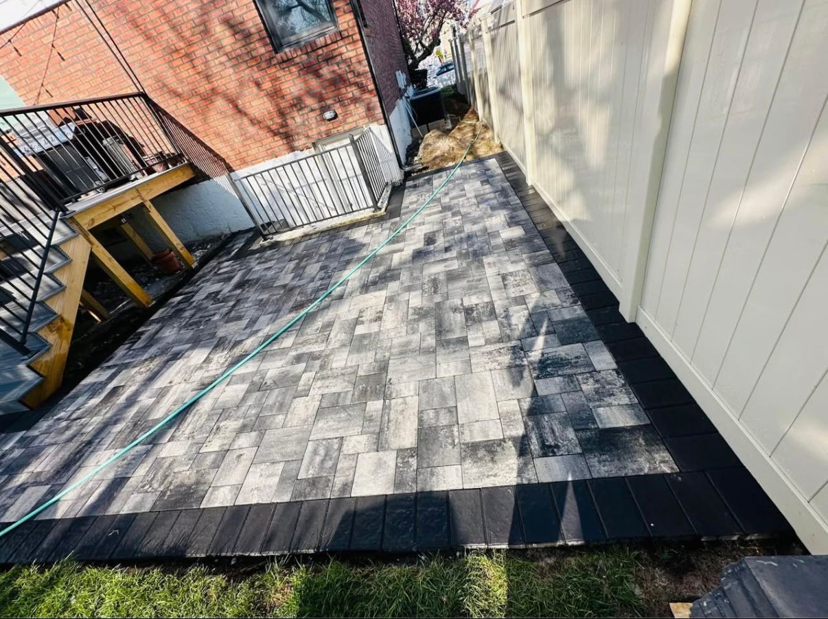 Overhead view of a backyard patio with gray pavers and black border, next to a white fence and a brick building.
