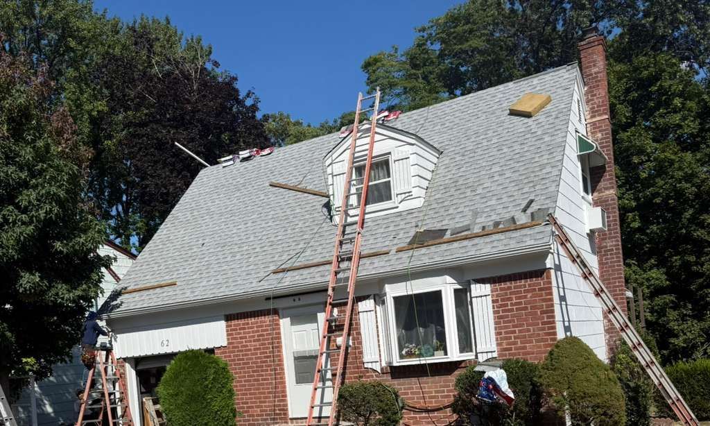 House with gray shingles under construction, ladders propped against it, brick exterior, blue sky.