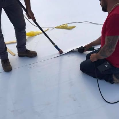 Two workers heat-seal a white membrane roof. One kneels, the other stands, both using tools.