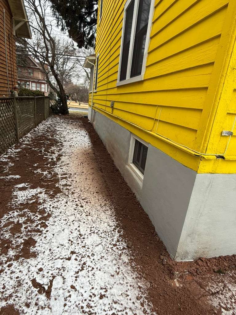 Yellow house side with a gravel bed along the foundation, snow patches, and a small window.