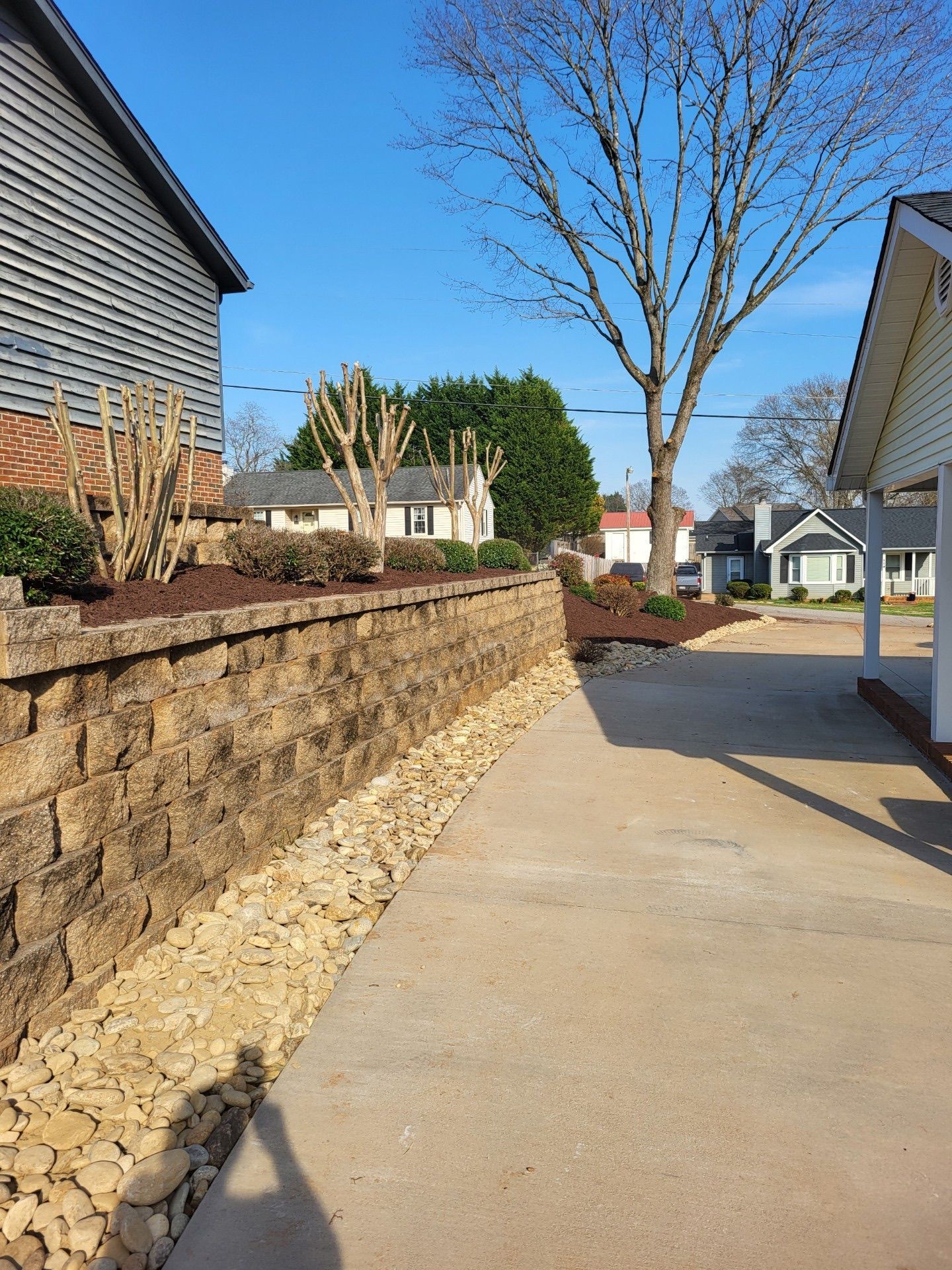 Sunlit concrete sidewalk beside a stone retaining wall and houses under a clear blue sky.