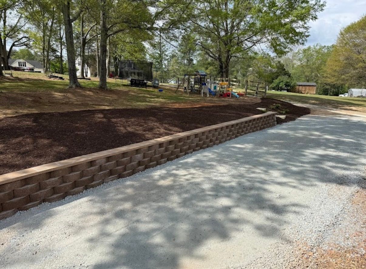 Gravel path beside a curved brick retaining wall in a wooded park with playground equipment in the background