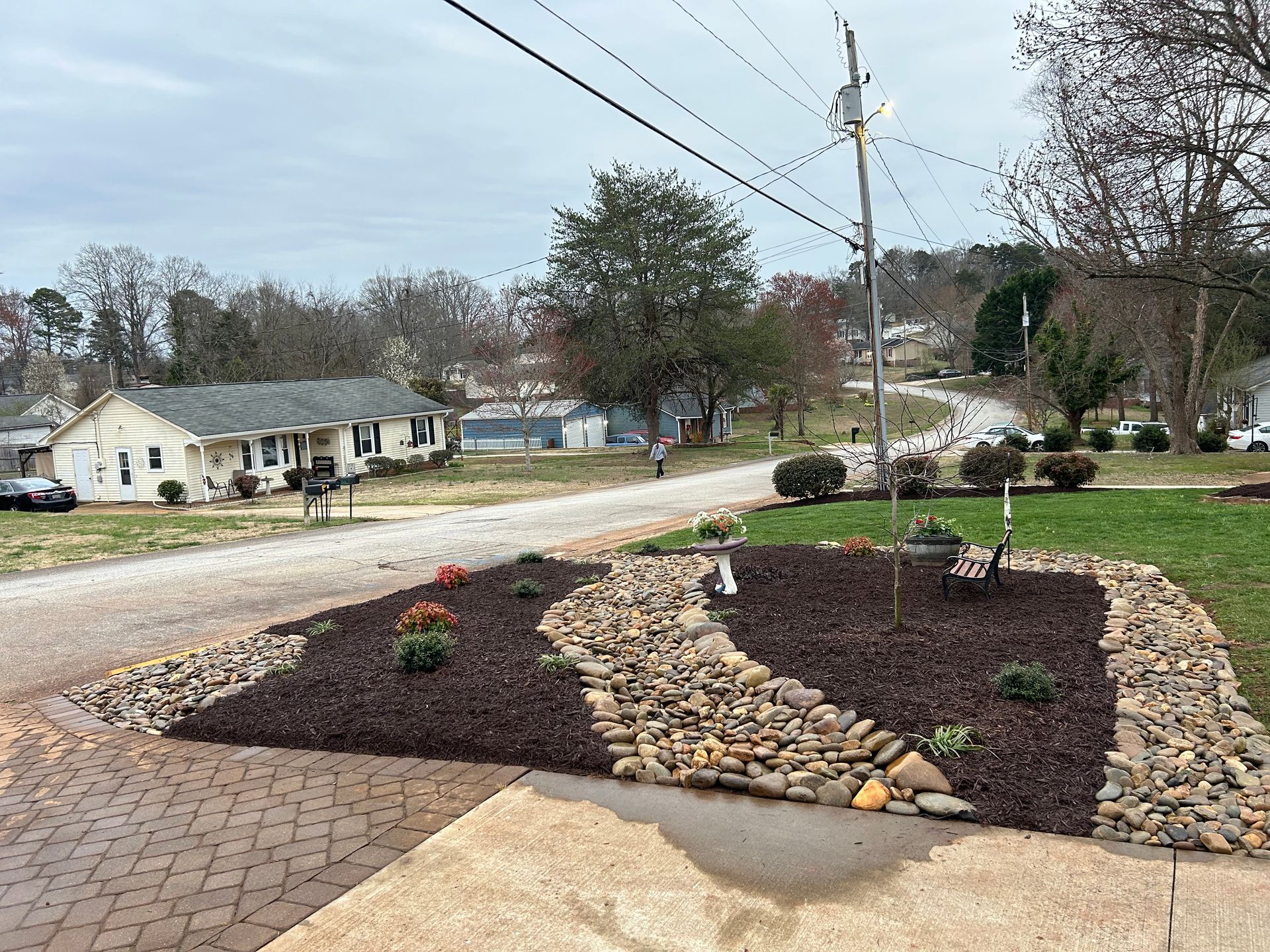 Landscaped front yard with mulch beds, rocks, small plants, and a brick driveway beside a quiet suburban street