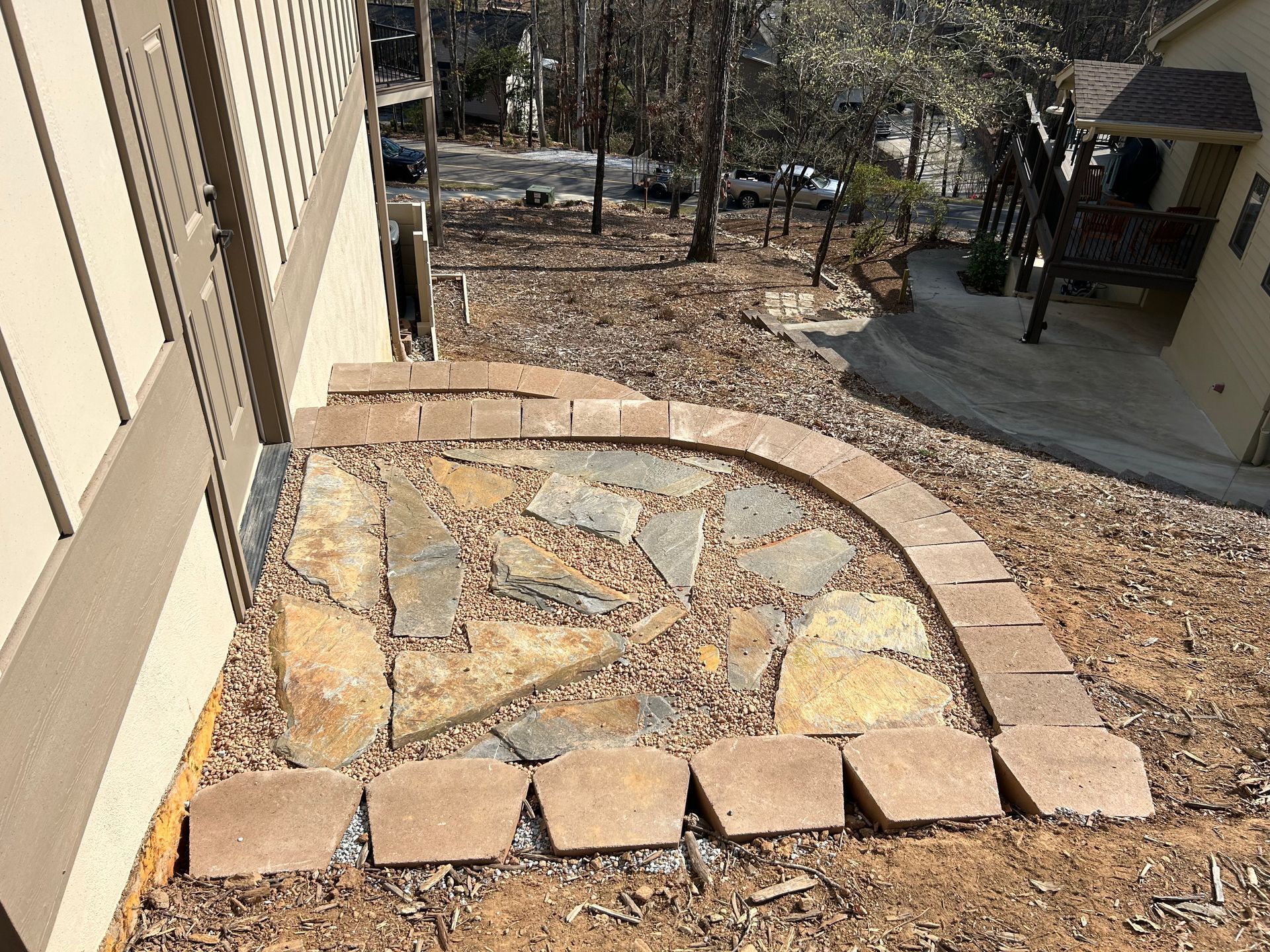 Stone mosaic patio beside beige house with curved brick border overlooking a wooded yard