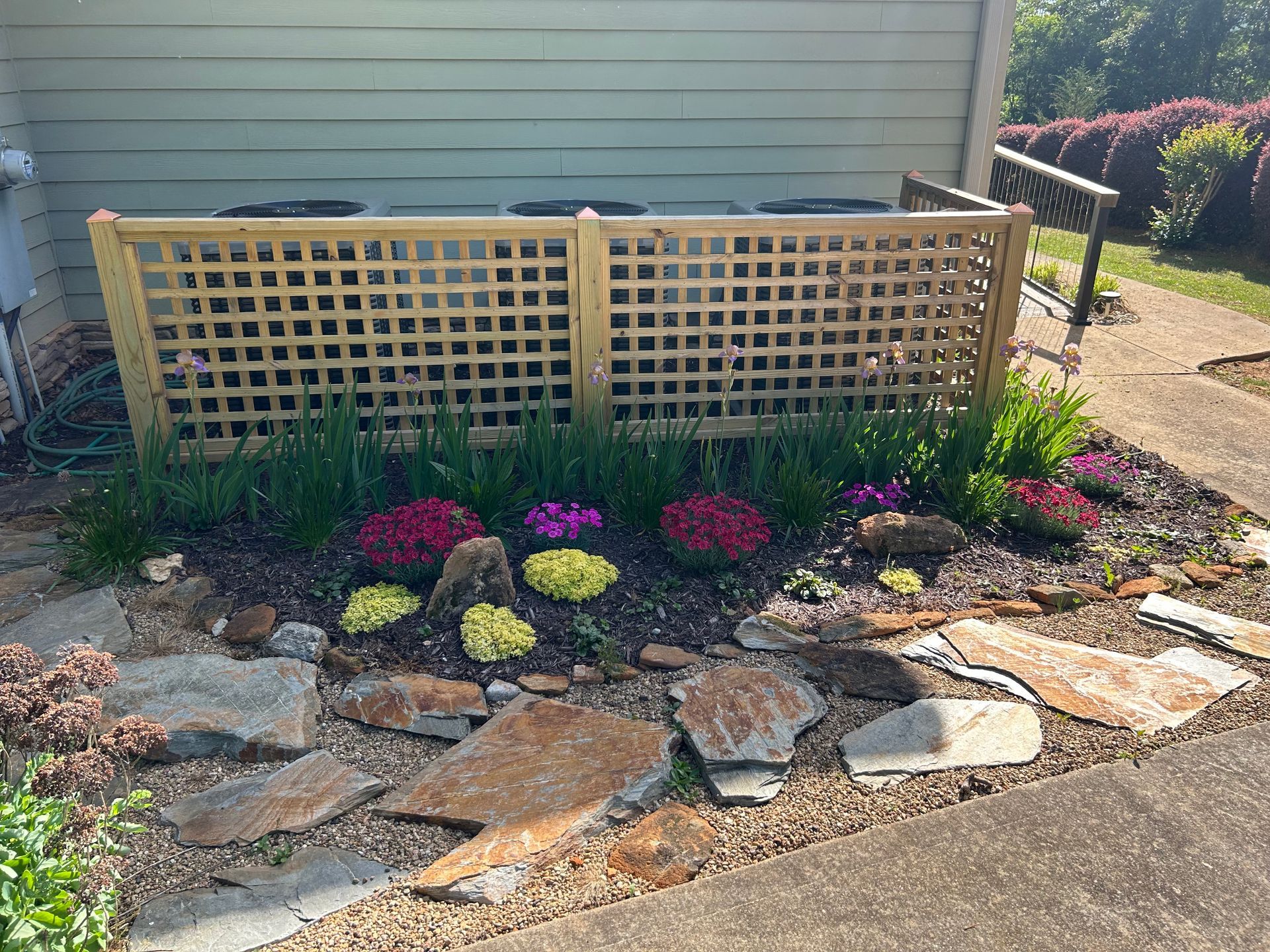 Garden bed with a wooden lattice fence, colorful flowers, stone edging, and a house wall in the background