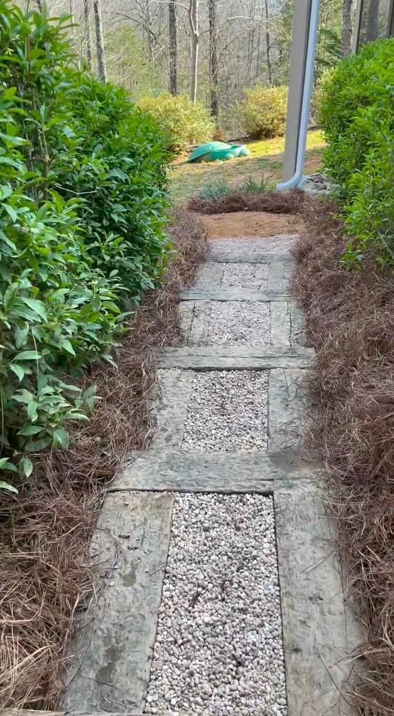 Narrow stone garden path between hedges, leading downhill to a yard with a wheelbarrow and yellow tarp.