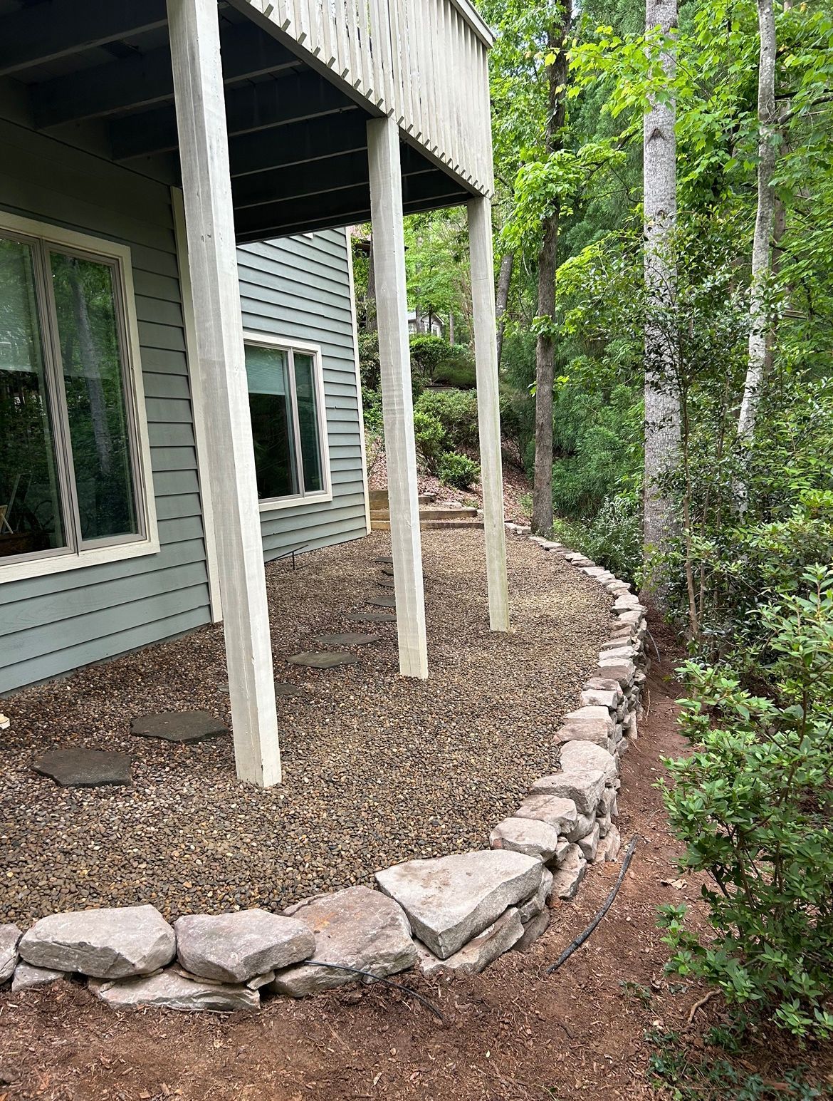 House exterior with a covered porch, gravel landscaping, and a stone border beside dense green trees