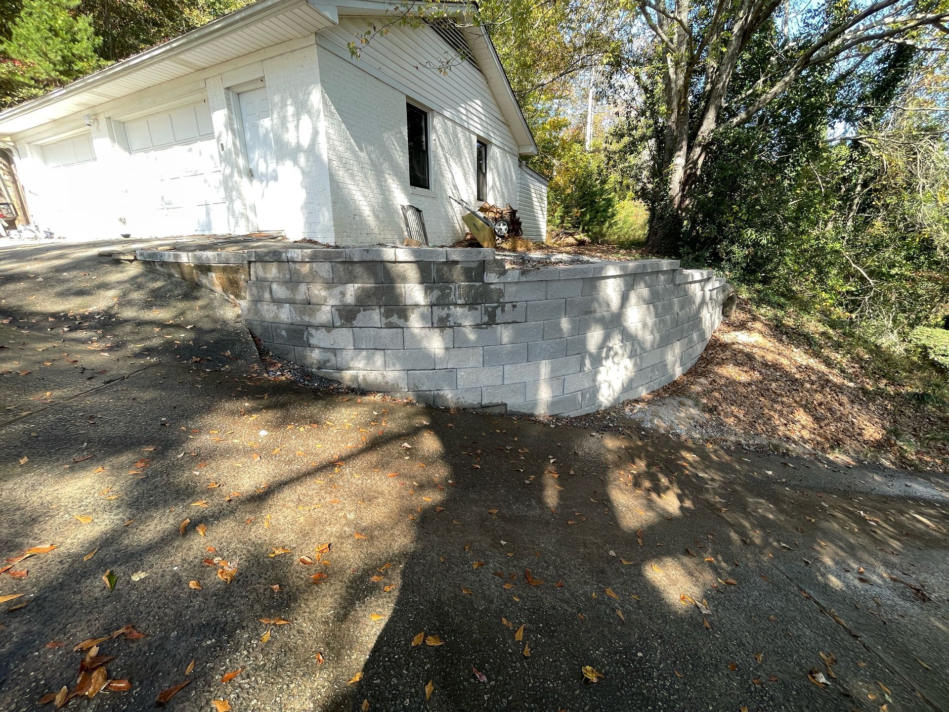 White house beside a curved stone retaining wall on a shaded wooded slope