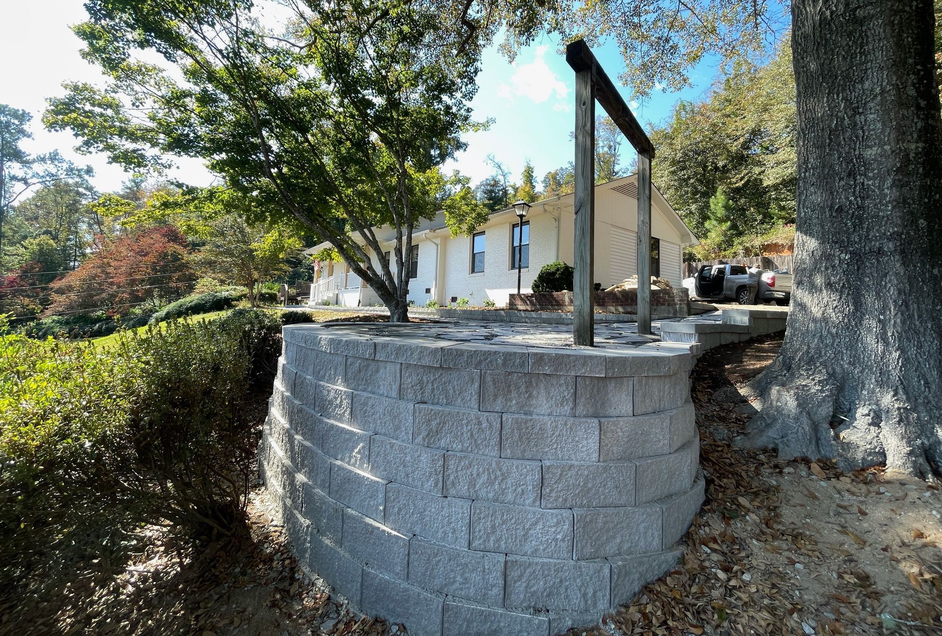 Stone retaining wall with wooden handrail beside a tree-lined path and house in the background