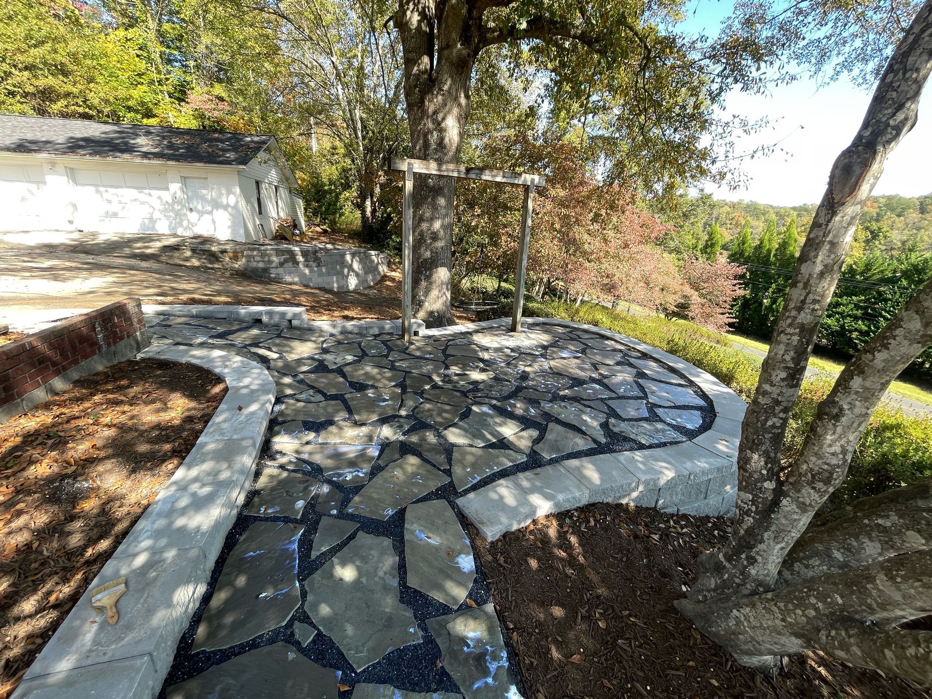 Stone garden path leading to a wooden arbor, lined with trees and bordered by mulch in a sunny yard