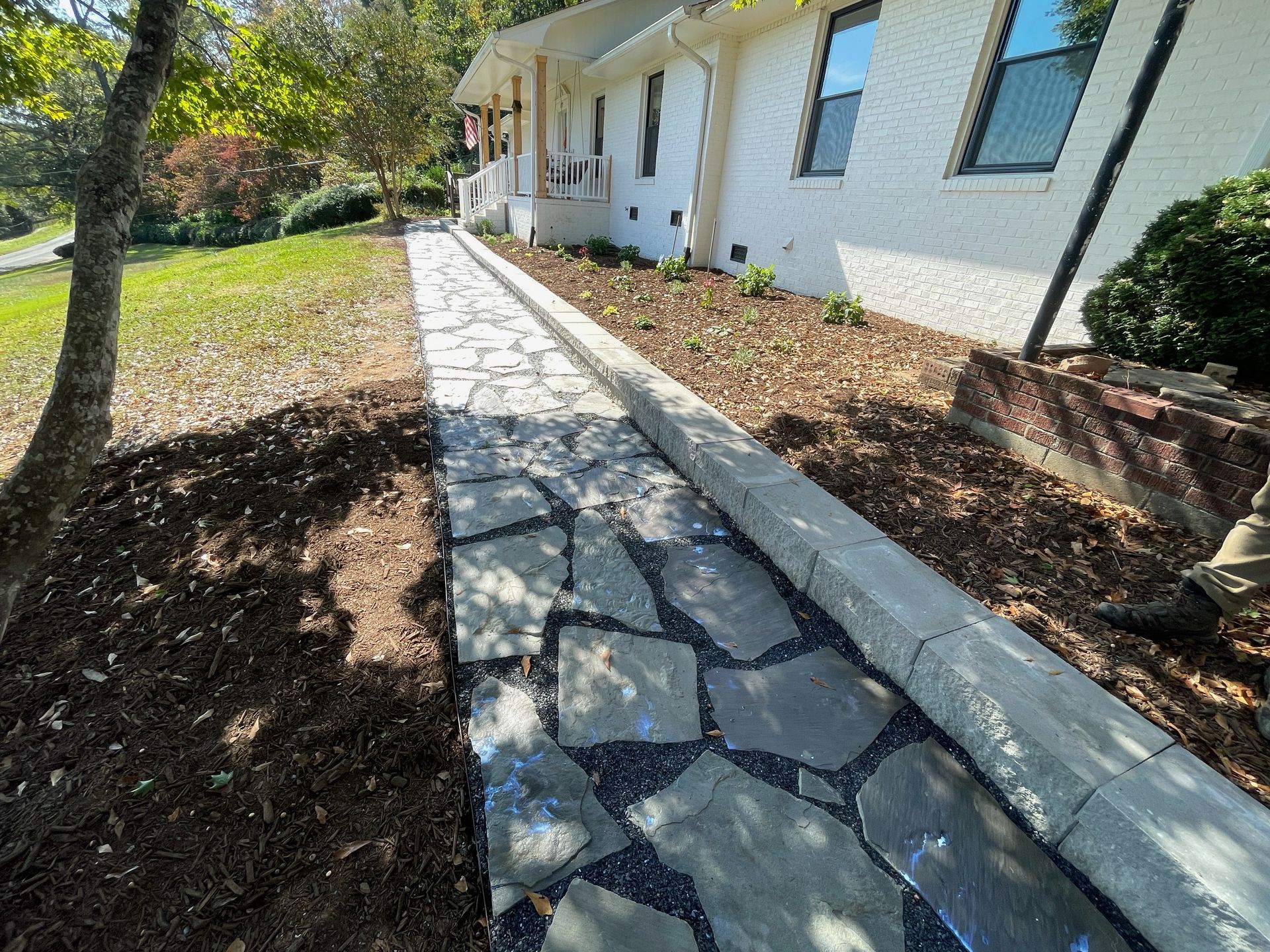 Stone walkway beside a white house, bordered by mulch and shrubs in a sunny yard.