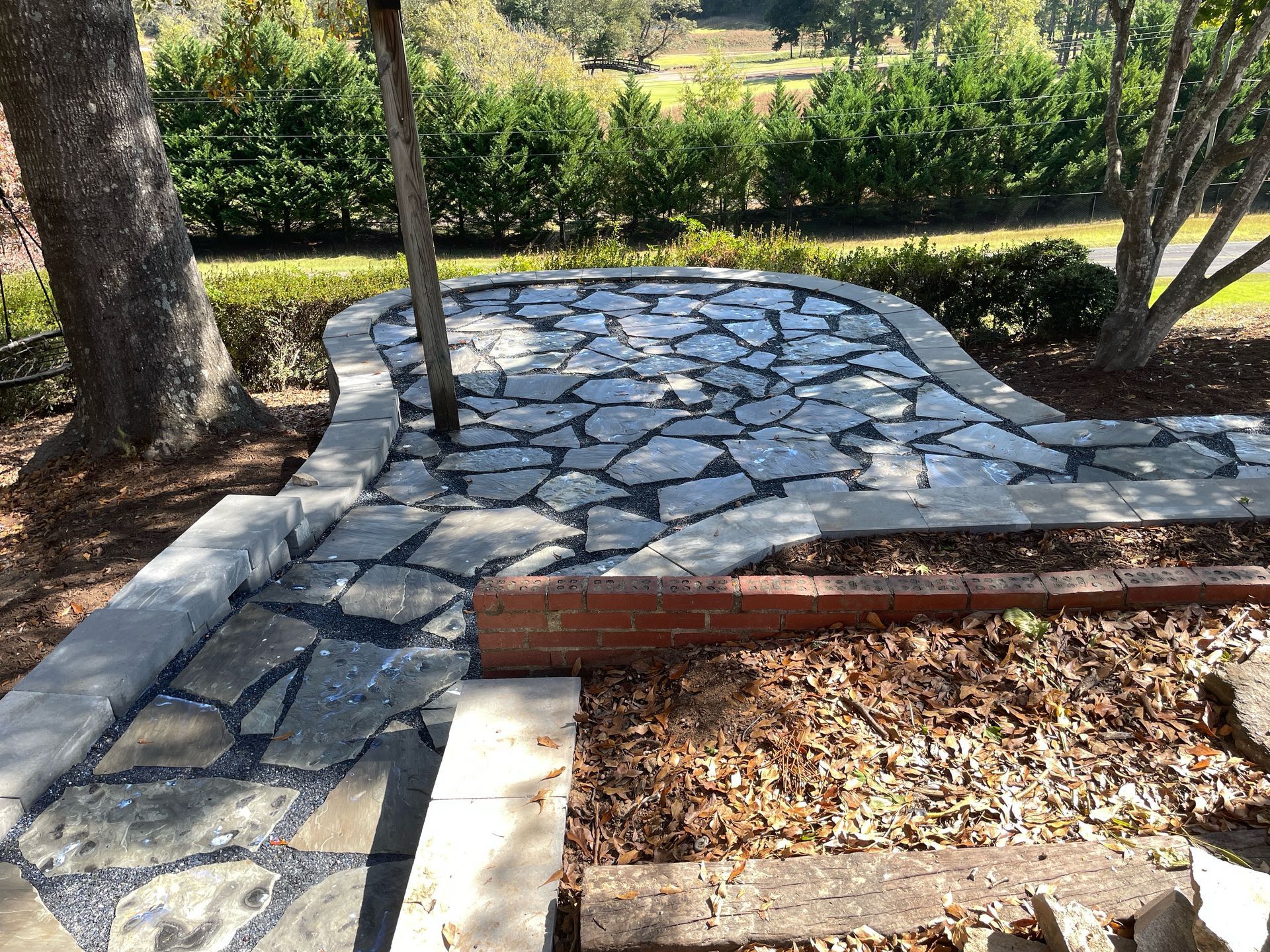 Stone garden path bordered by mulch and trees, with a wooden beam and greenery in sunlight