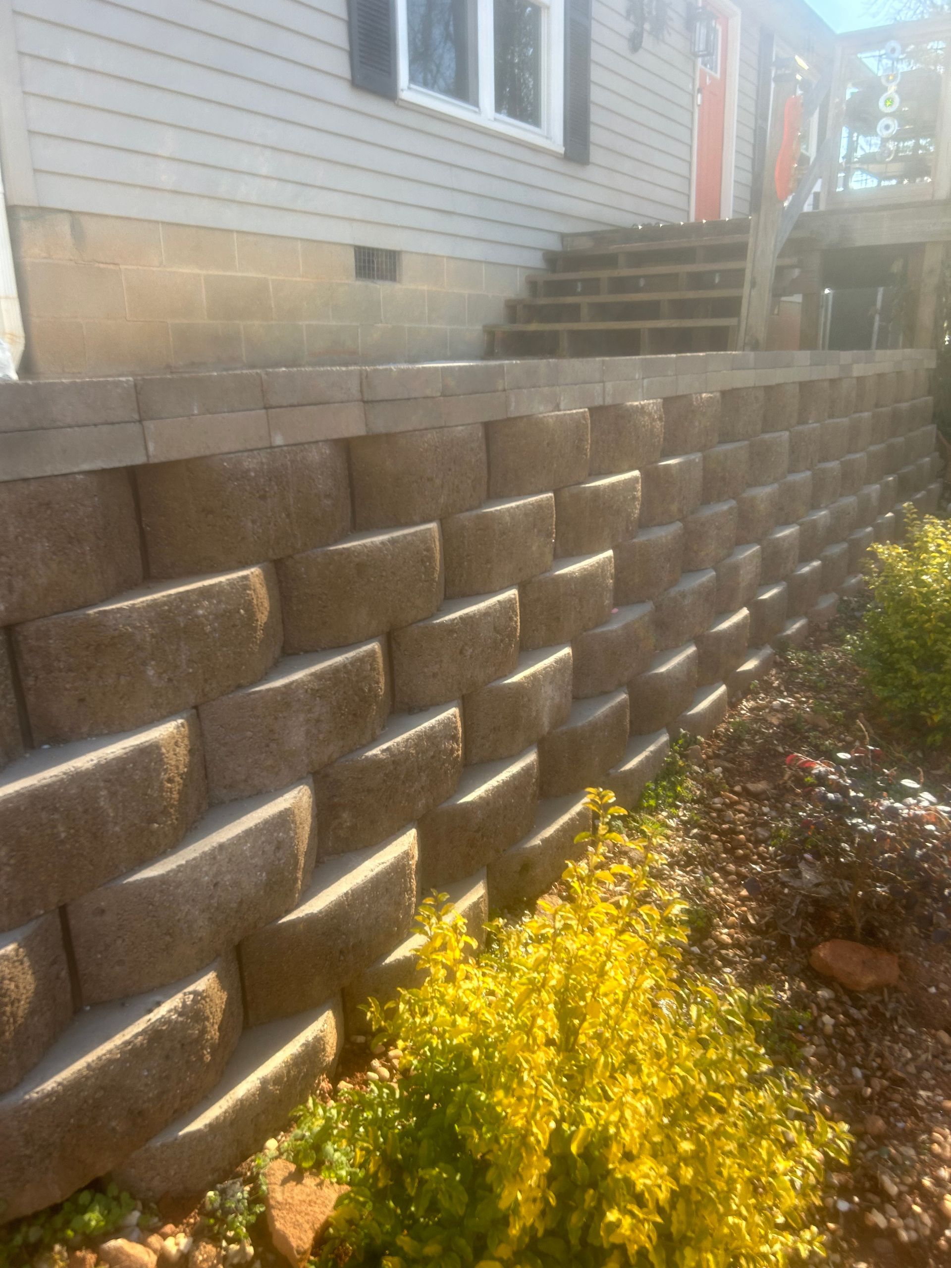 Stone retaining wall beneath house siding with yellow shrubs in the foreground and stairs at the top