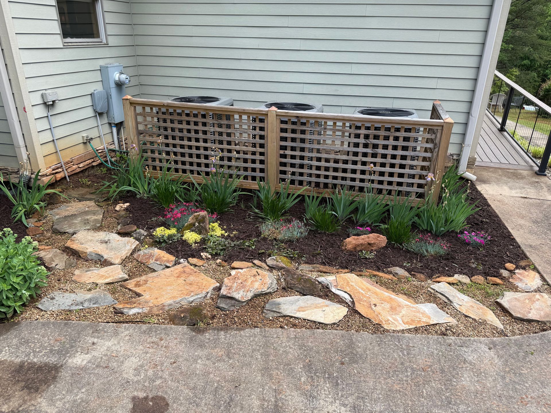 Small garden bed with stone border and trellis against a house wall beside a walkway.