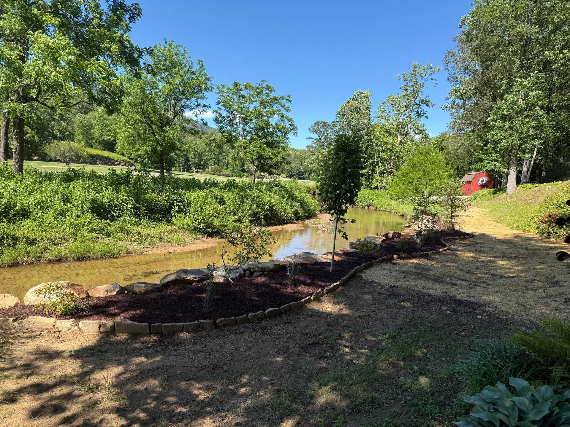 Sunny garden with a narrow stream, flower beds, trees, and a small red shed in the background
