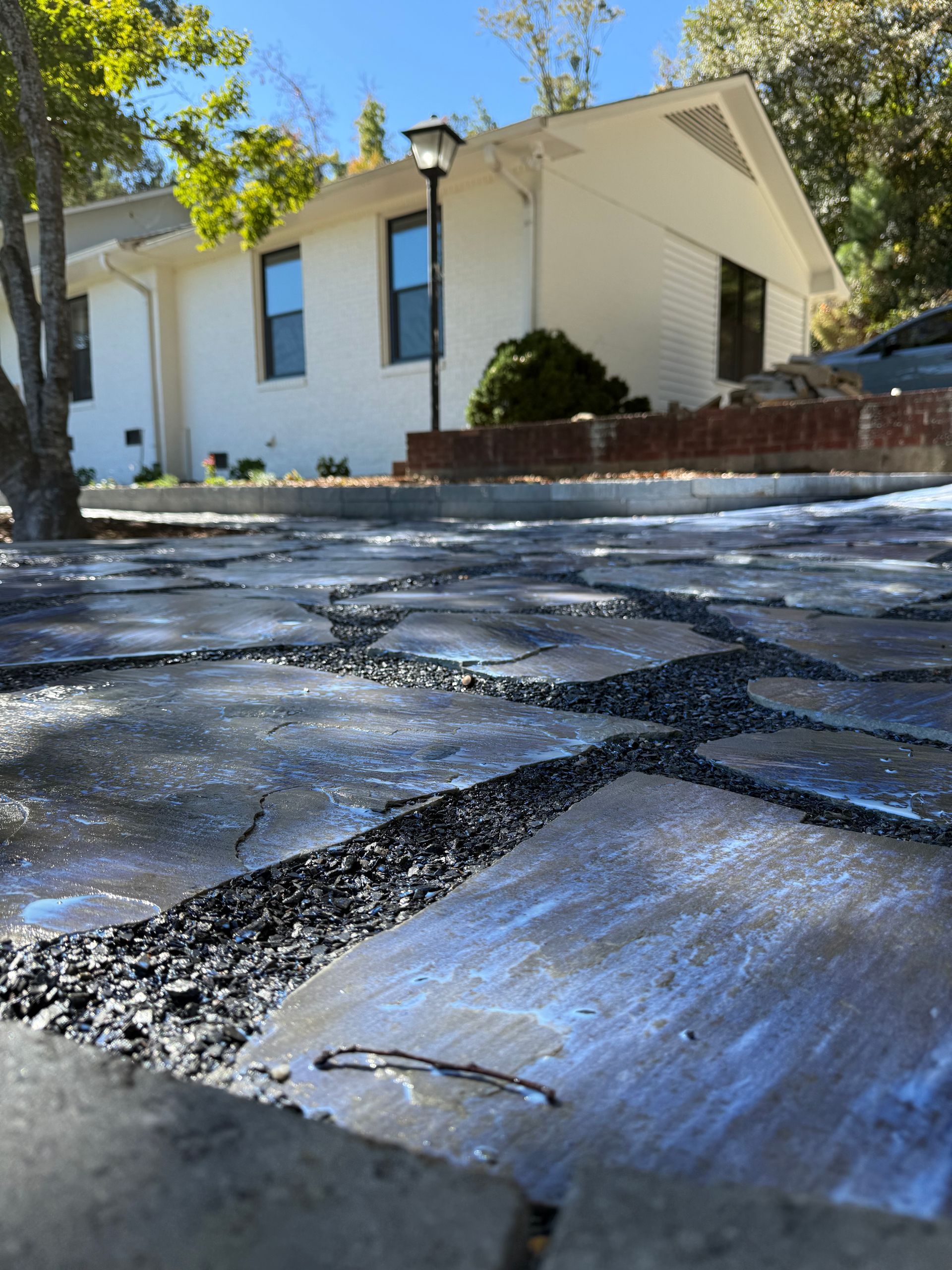 Cracked stone walkway in front of a white house with a brick planter and trees in bright sunlight