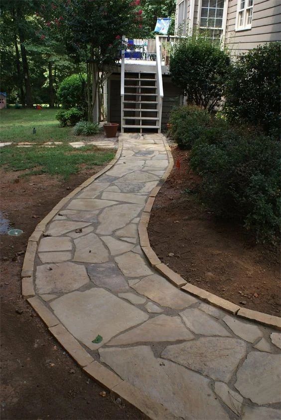 Curved stone walkway leading to house steps, bordered by shrubs and dirt yard