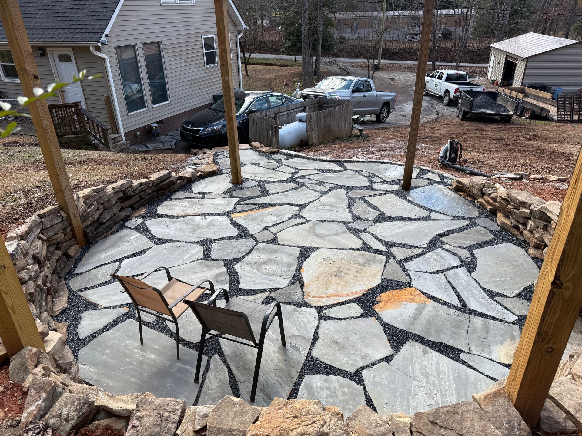 Stone patio in a backyard under construction, with chairs and wooden posts in front of a house.