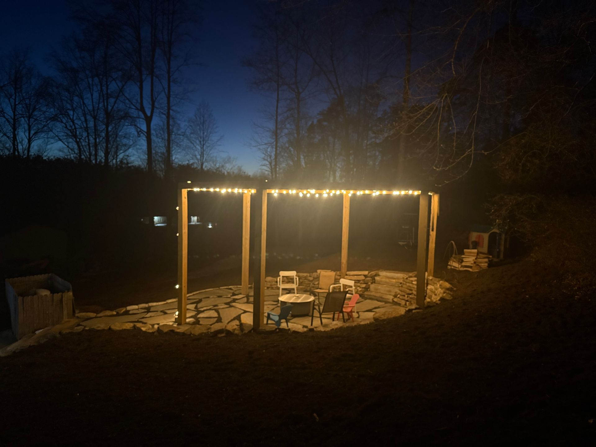 Lit outdoor patio with pergola, table and chairs at dusk in a wooded yard