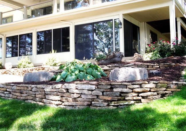 House with large windows and a stone retaining wall in a sunny landscaped yard