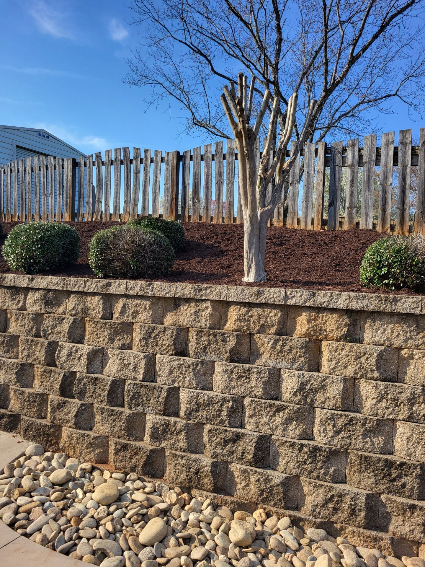 Stone retaining wall with shrubs, a young tree, and a wooden fence under a blue sky