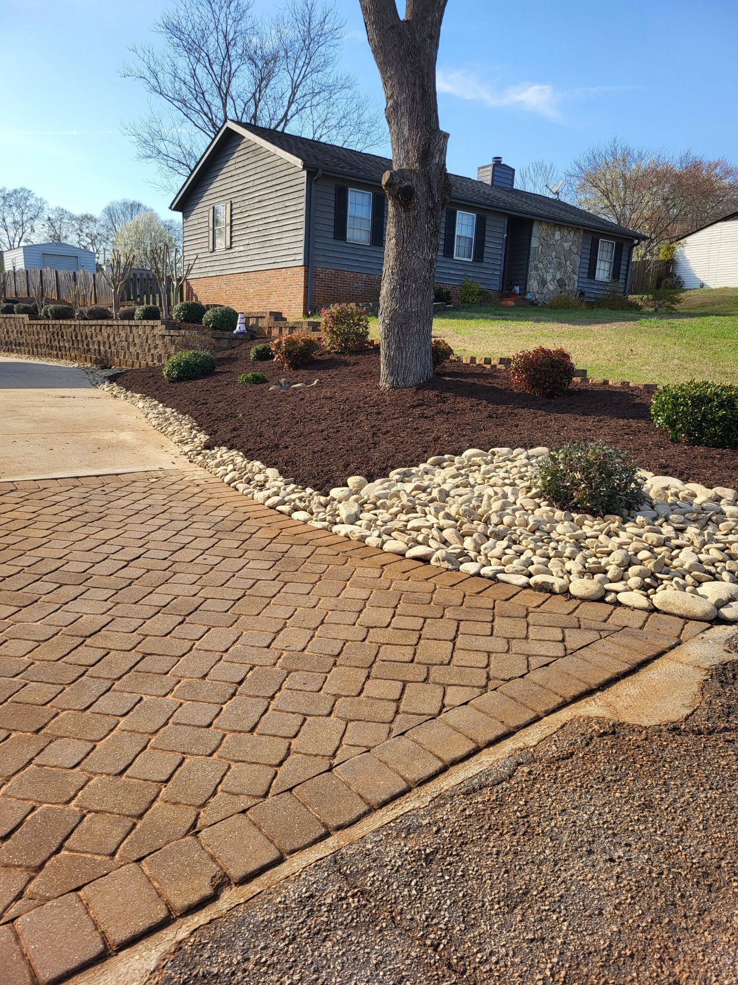 Brick walkway leading to a house with landscaped beds, trees, and a clear blue sky