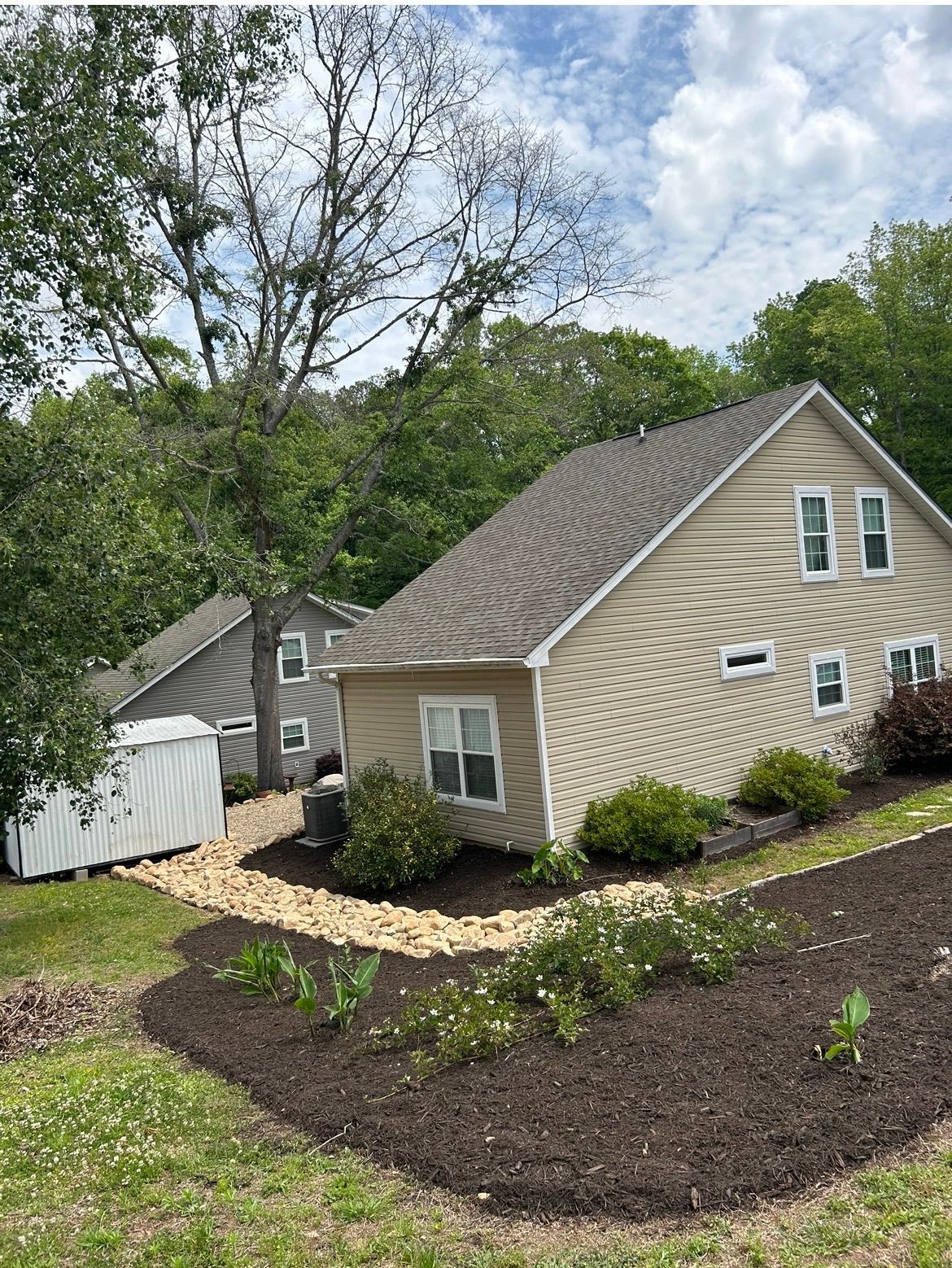 Beige house with white trim on a sloped landscaped yard, with trees and cloudy sky behind it