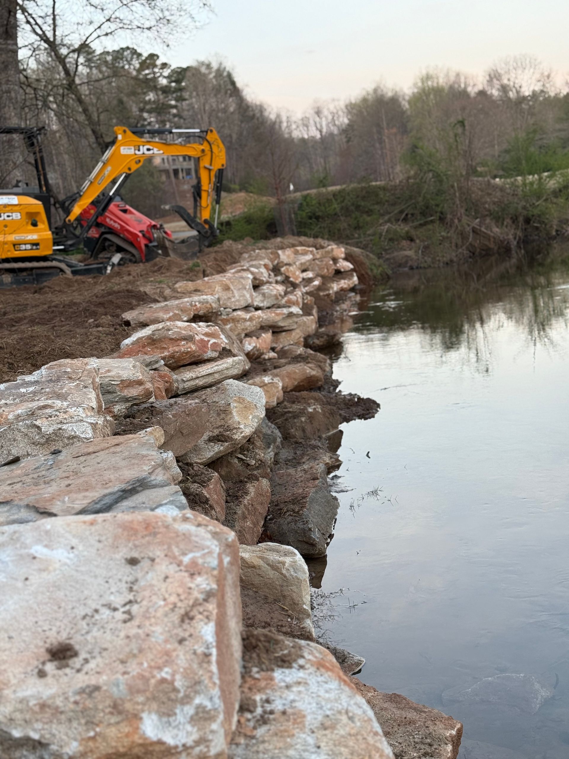 Excavator building a stone embankment beside a calm river in a wooded area.