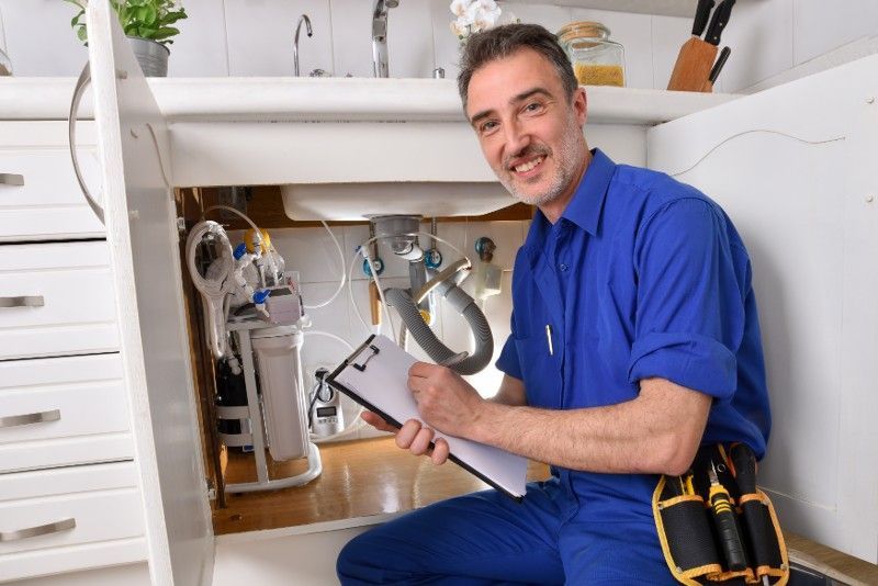 A plumber is kneeling under a sink holding a clipboard and smiling.