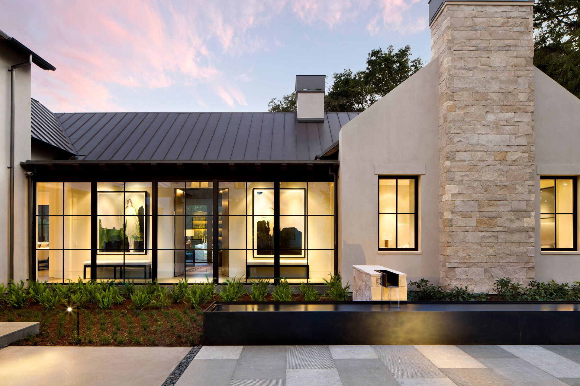 Modern stone-and-glass house exterior at dusk with warm interior lights and landscaped front yard