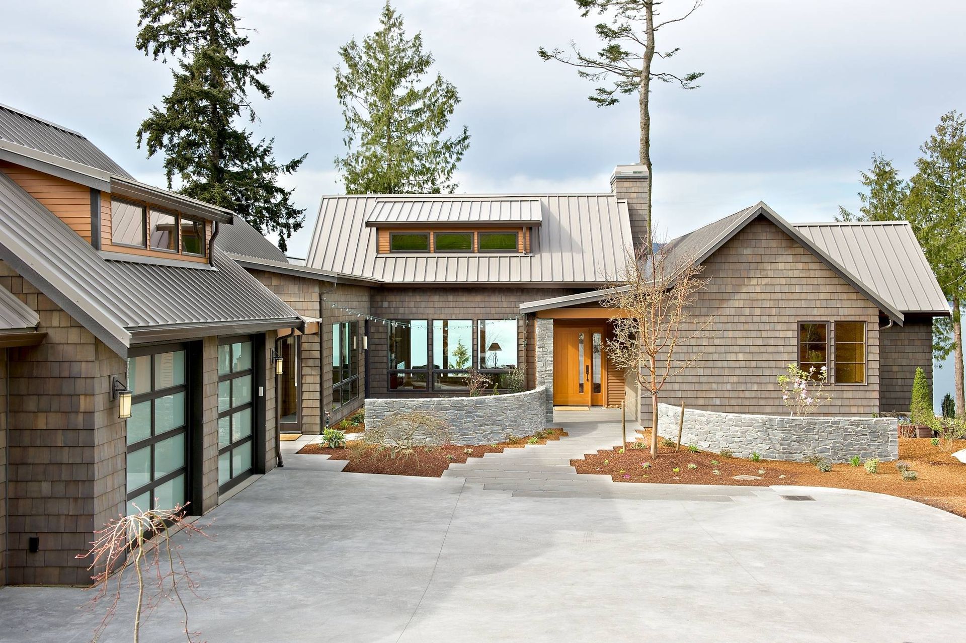 Modern rustic house exterior with stone and wood siding, garage, and curved driveway surrounded by trees