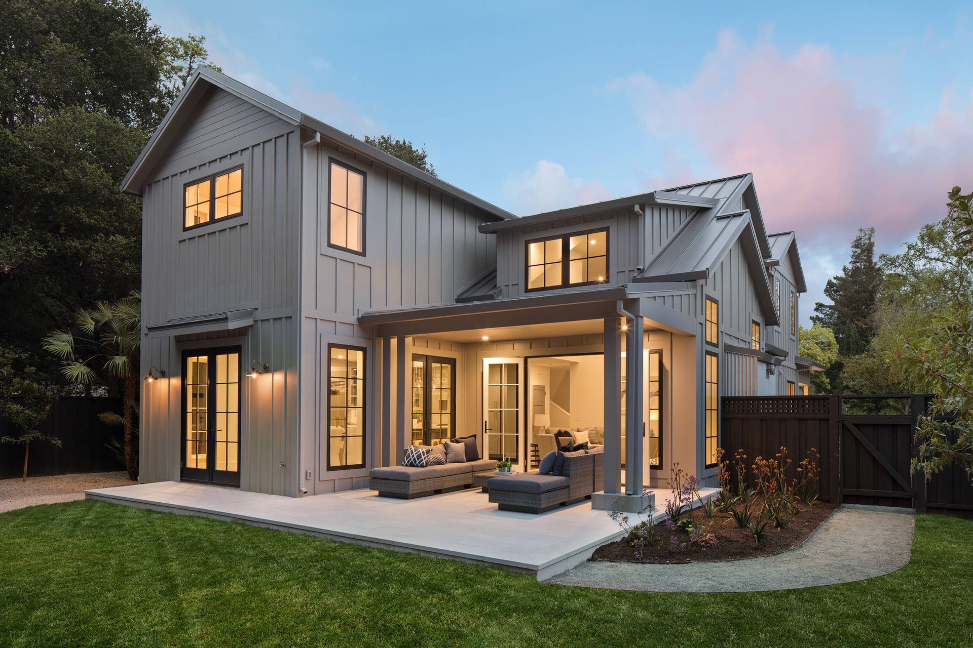 Modern two-story gray house at dusk with warm interior lights, patio seating, and a lit backyard