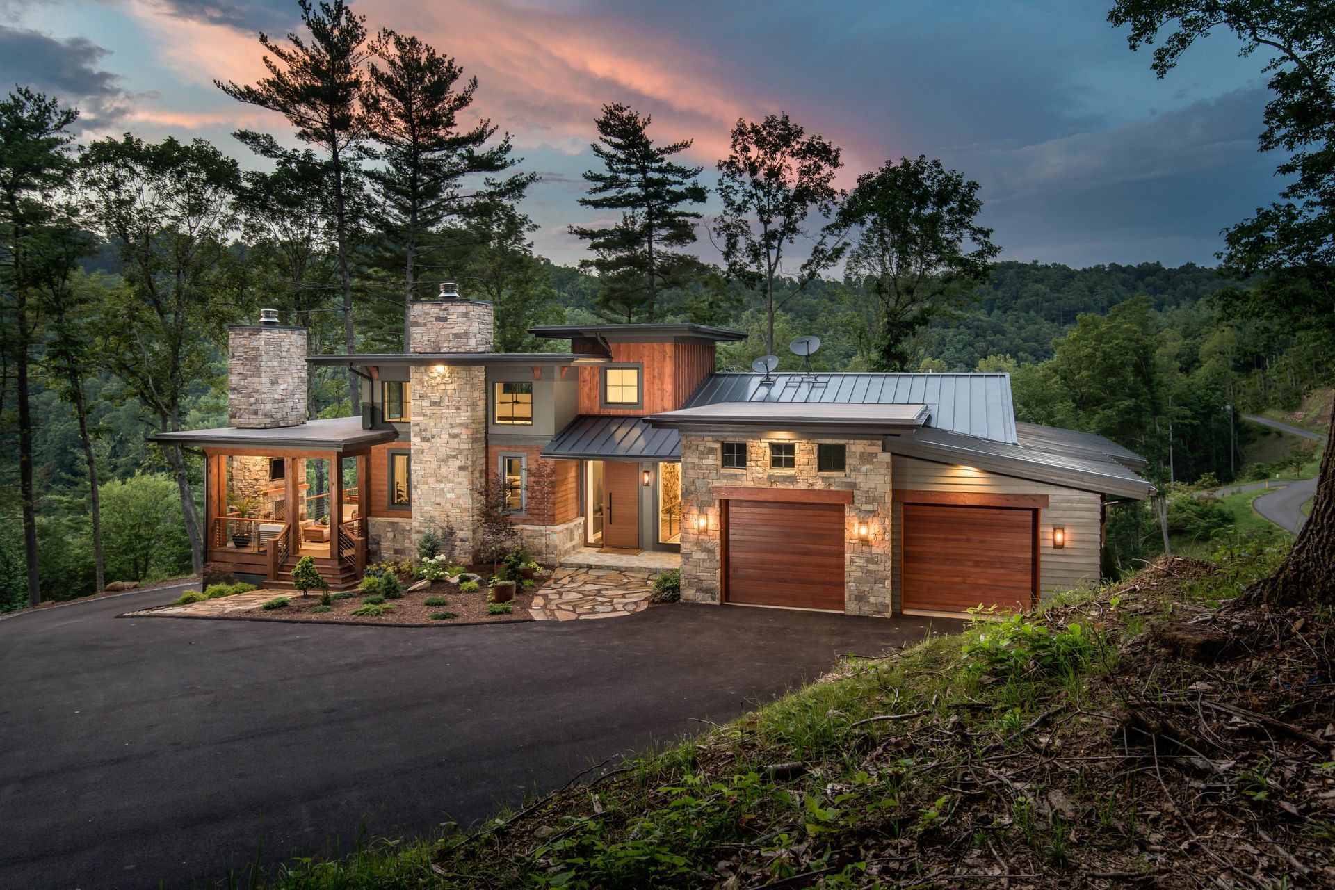 Modern stone-and-wood house at dusk with lit windows, garage, and forested hillside backdrop