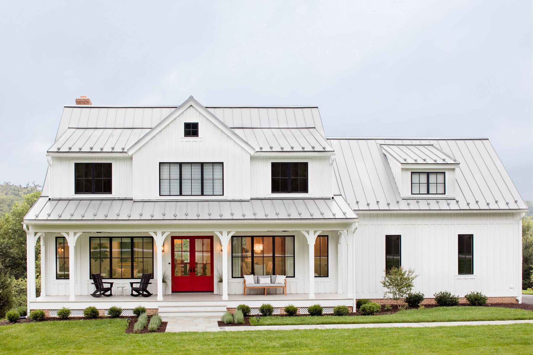 White farmhouse with red front door, metal roof, and covered porch on a green lawn