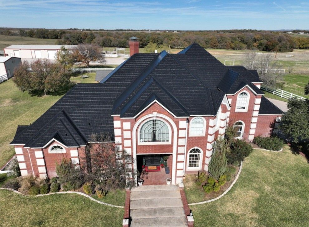 Aerial view of a large red-brick house with a dark roof and front walkway in a rural setting
