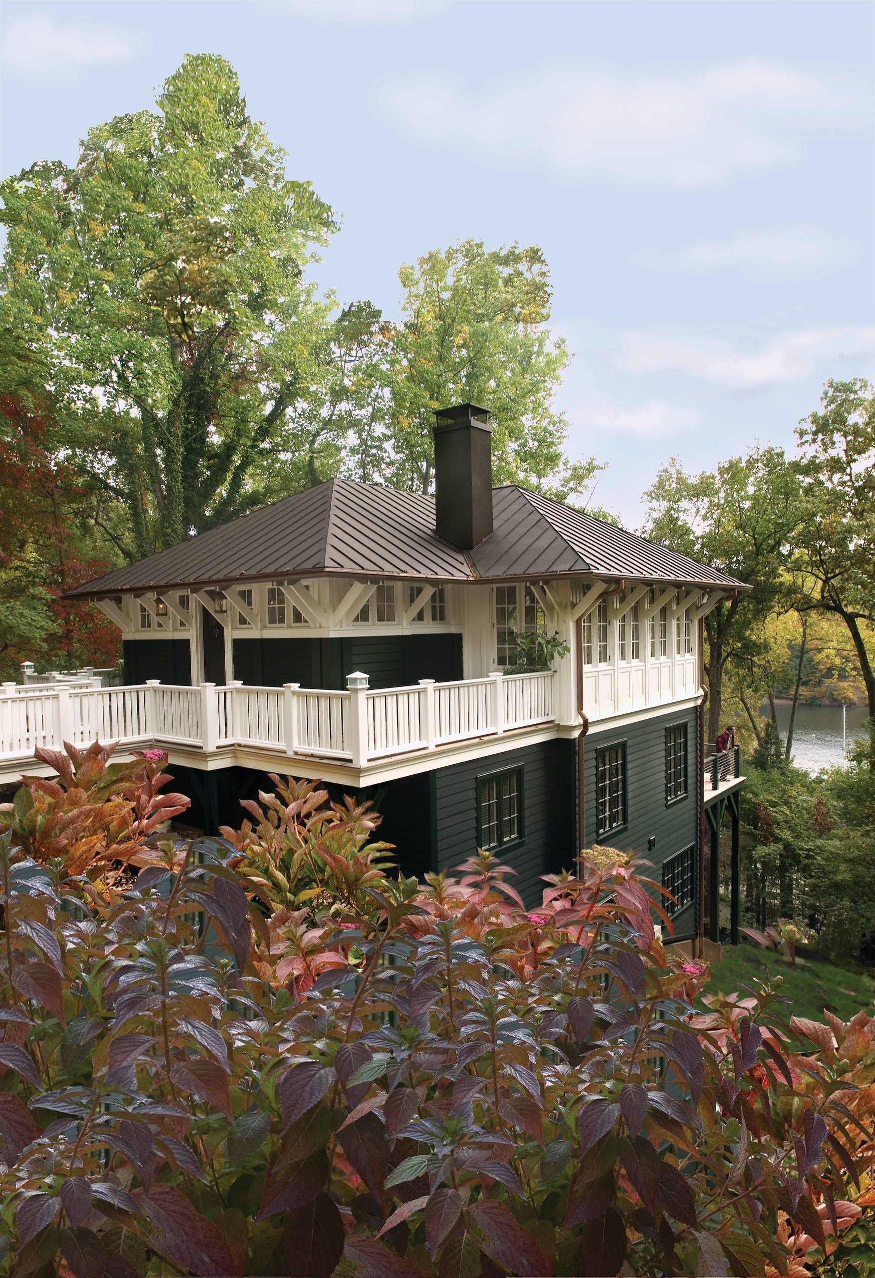 Two-story white house with a black roof and porch, surrounded by lush green trees and shrubs.