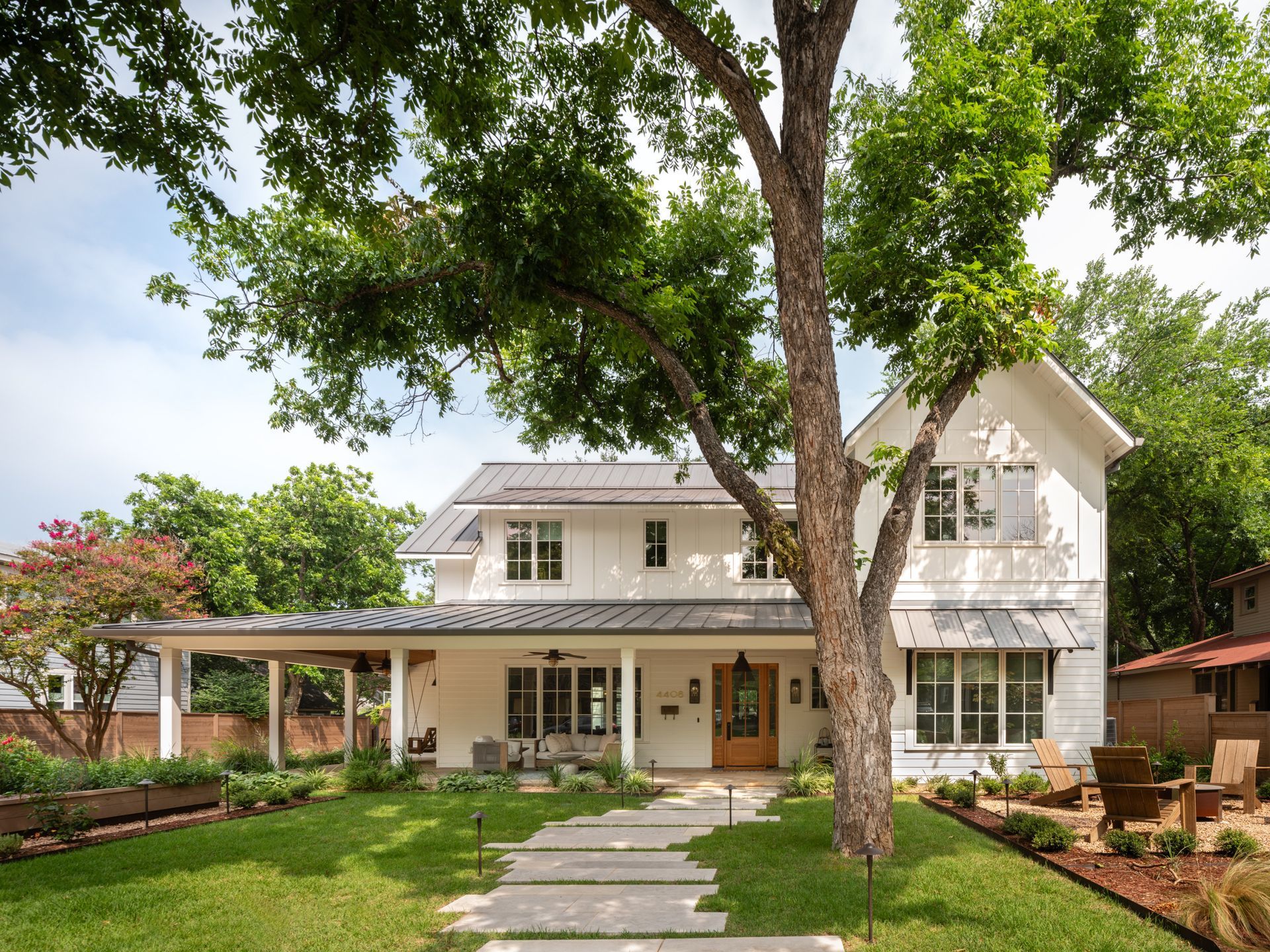 White two-story house with a front porch and stone walkway, framed by a large tree and green lawn