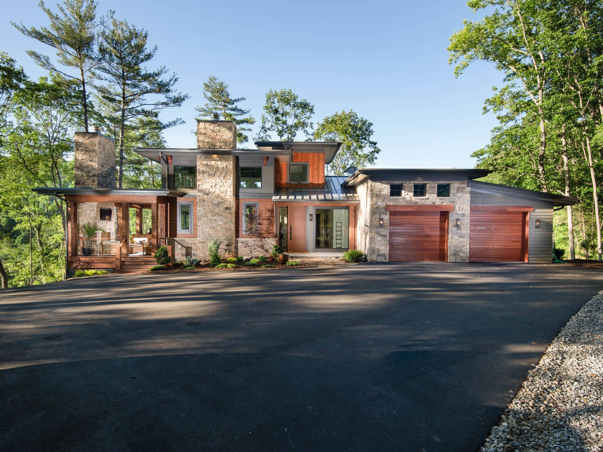 Modern two-story house with stone and wood exterior, large driveway, and surrounding trees under a clear sky