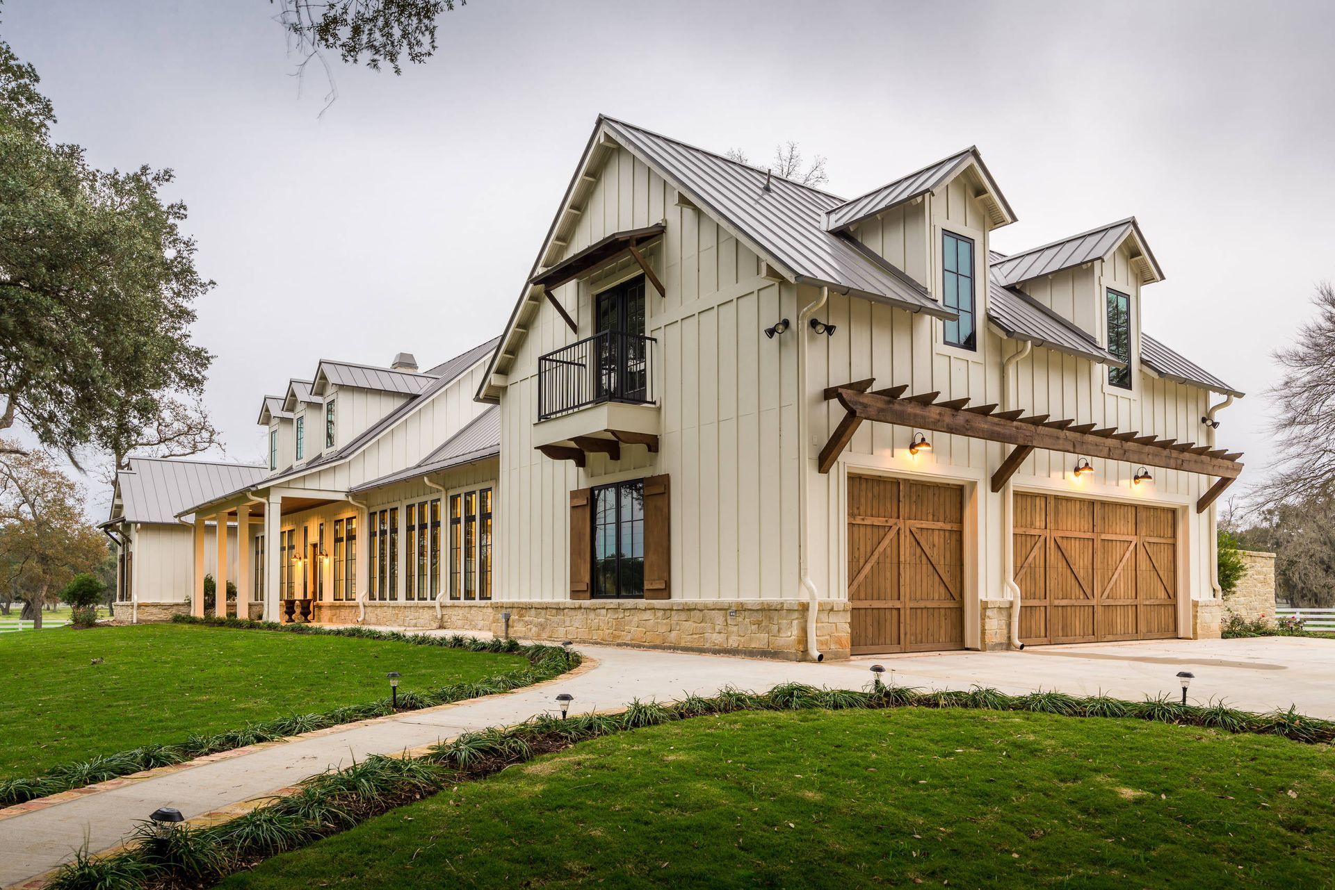 Large white barn-style house with a metal roof, wooden garage doors, and a green lawn under an overcast sky