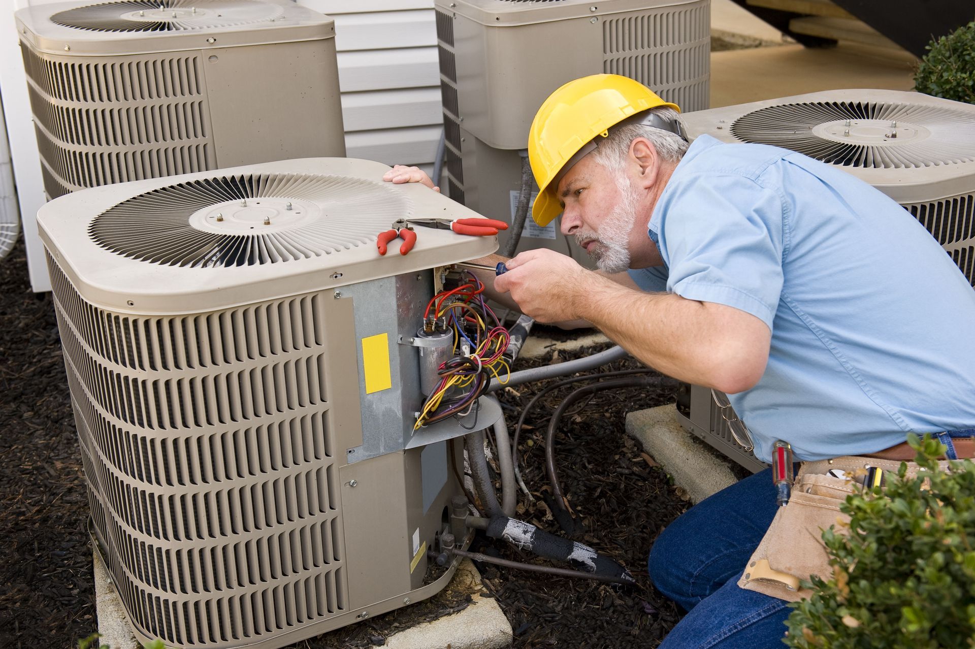 HVAC technician in a yellow hard hat repairs an air conditioner outside.