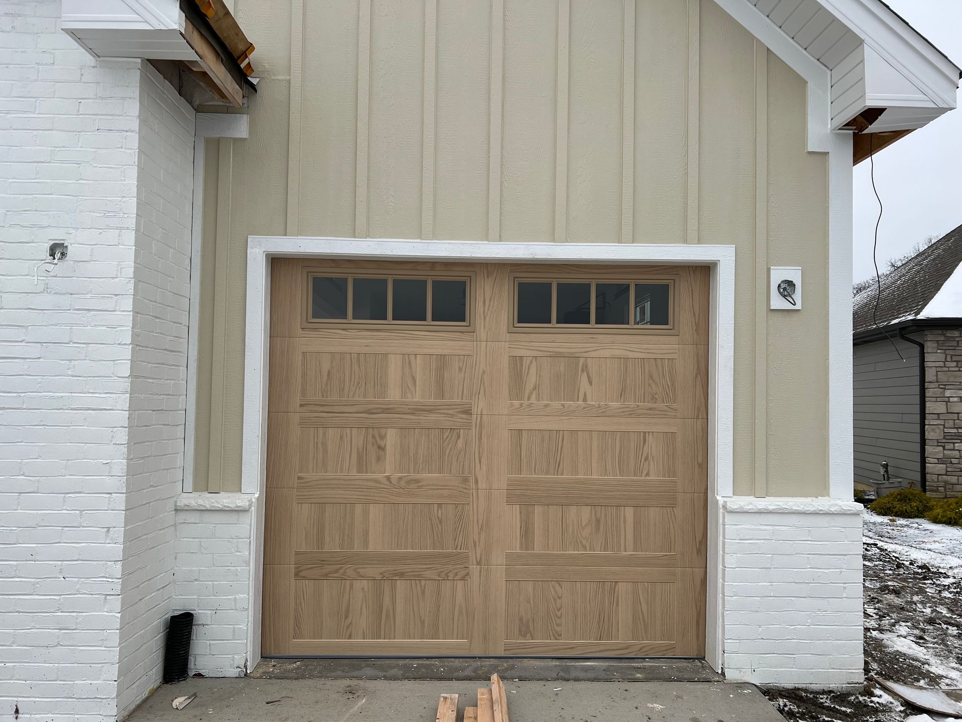 Light wood double garage door on a beige house exterior with white brick accents.