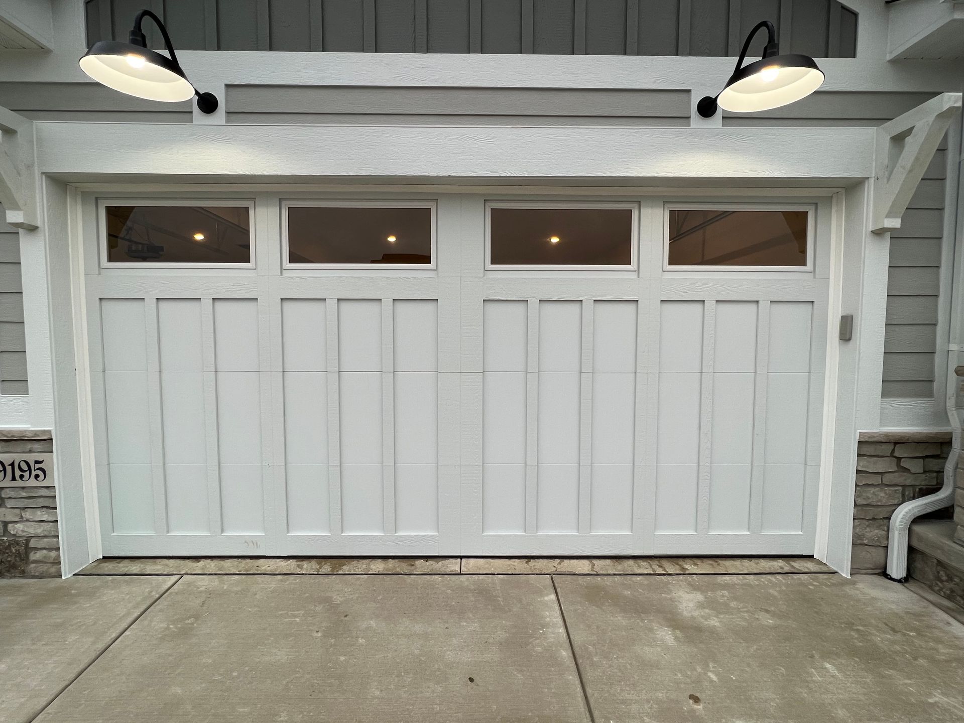 White garage door with four frosted windows, lit by two wall lamps on a driveway.