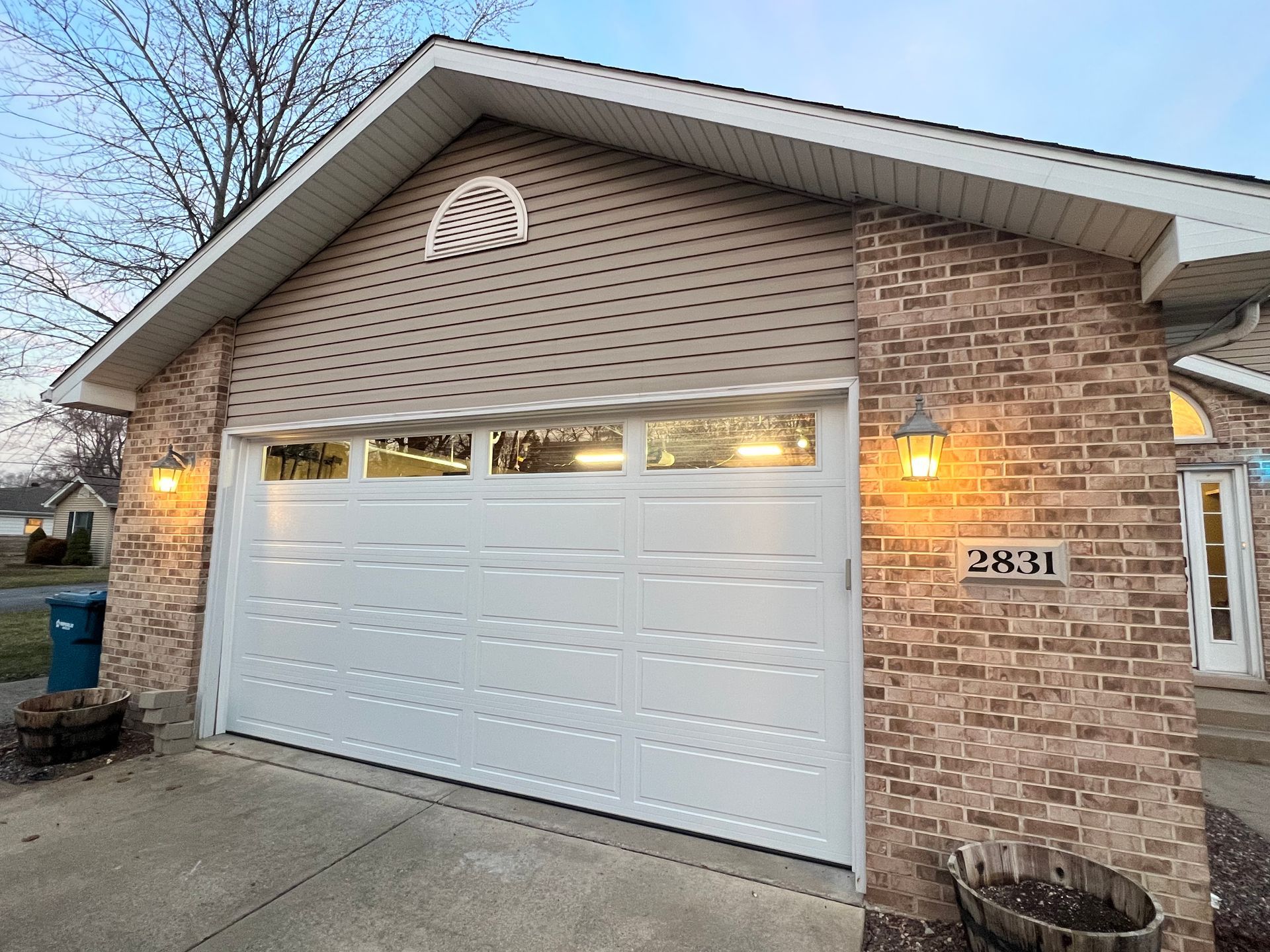 Brick house with a white garage door, lit porch lights, and concrete driveway at dusk